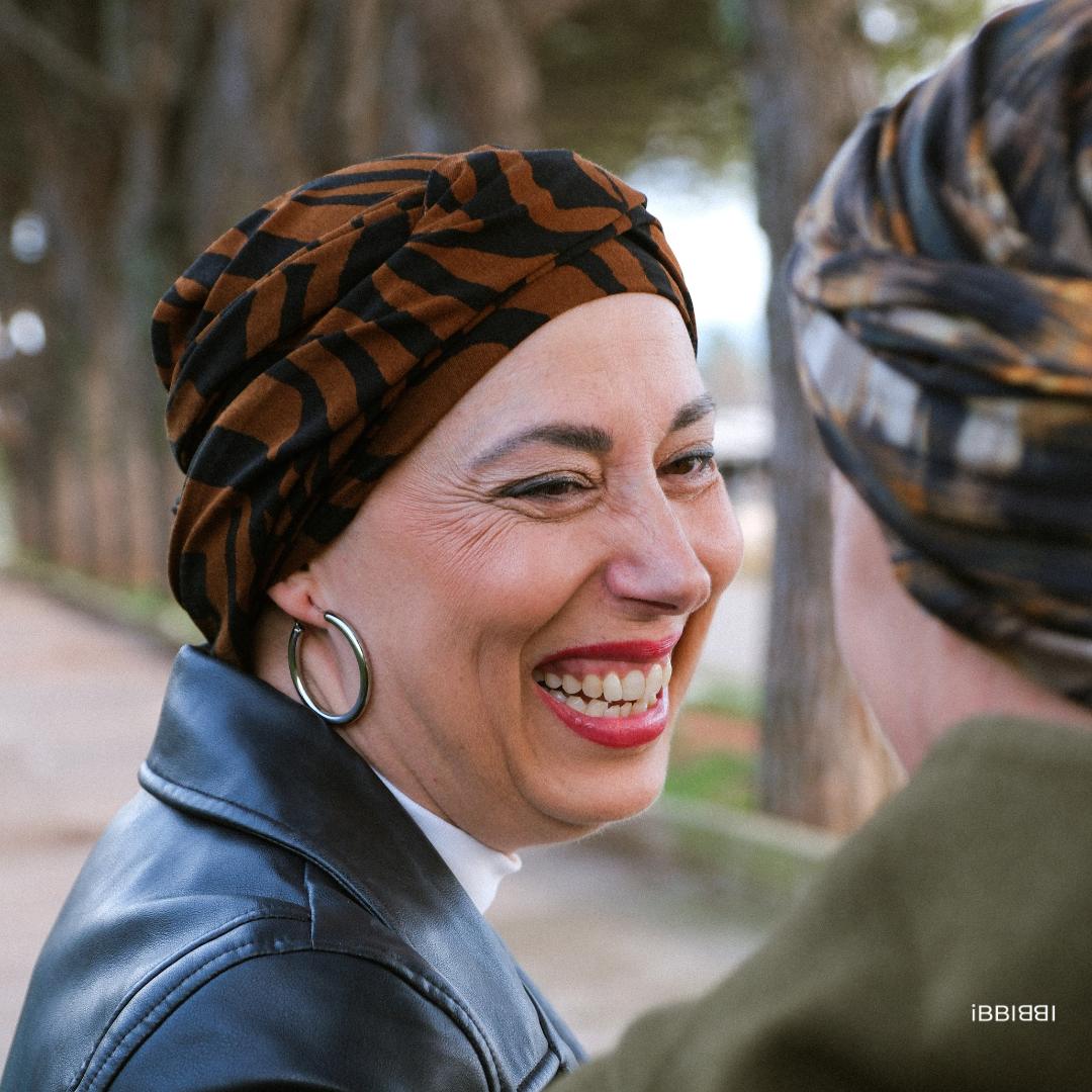 Mujer sonriendo con un pañuelo oncológico marrón con estampado negro de iBBIBBI, disfrutando de un momento al aire libre
