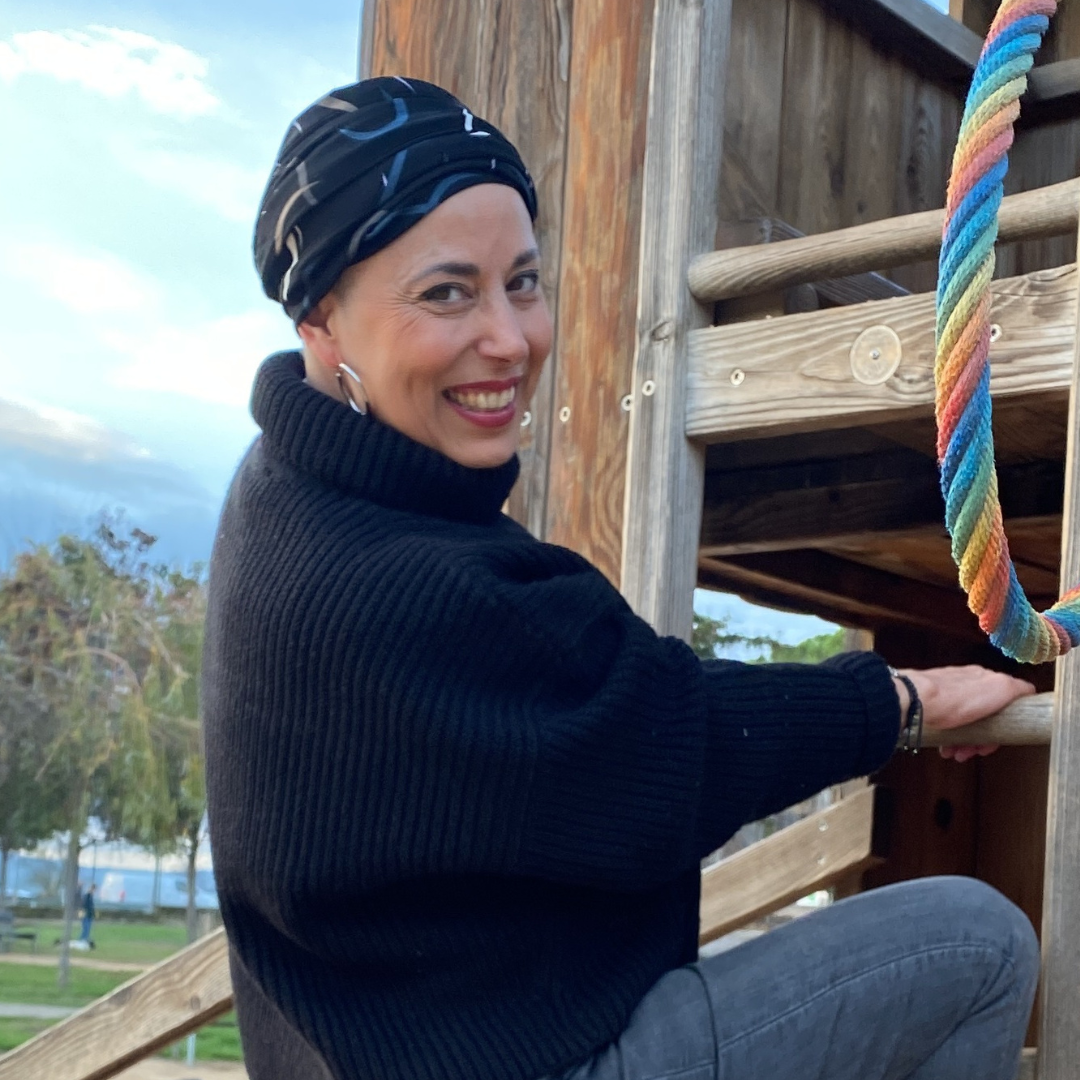 Mujer sonriente con un pañuelo negro oncológico modelo Savannah, disfrutando de un momento al aire libre. Lleva un suéter negro de cuello alto y posa junto a un juego de madera, transmitiendo comodidad, diversión y confianza.