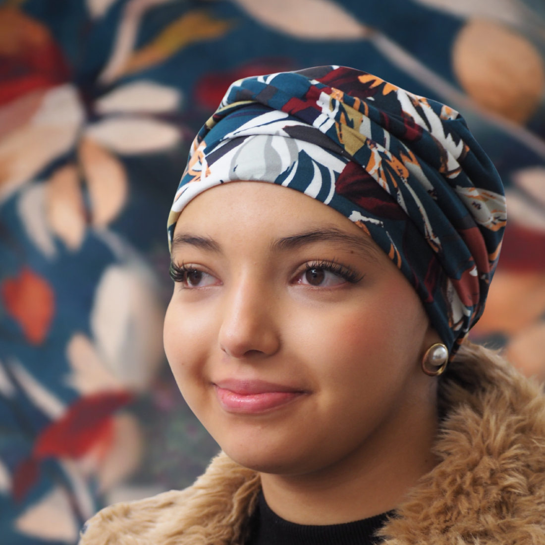 Mujer joven con turbante oncológico iBBIBBI estampado en tonos azules, teja y mostaza, mirando hacia abajo con una sonrisa suave. El fondo muestra el mismo estampado ampliado y desenfocado, creando una imagen armoniosa y serena.