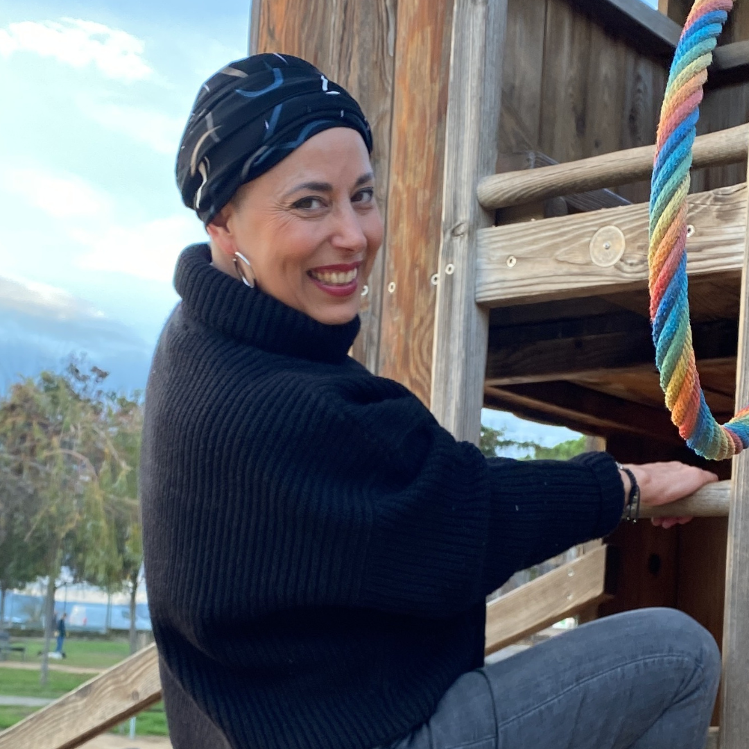 Mujer sonriente con un pañuelo negro oncológico modelo Savannah, disfrutando de un momento al aire libre. Lleva un suéter negro de cuello alto y posa junto a un juego de madera, transmitiendo comodidad, diversión y  confianza.